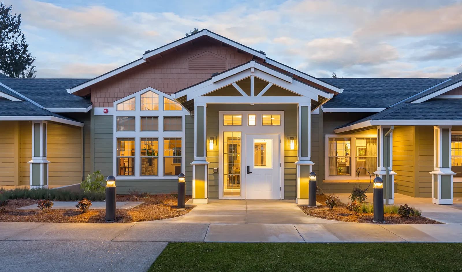 The entry of a craftsman-style building at dusk, lit by wall sconces, bollards along the path and warm interior light