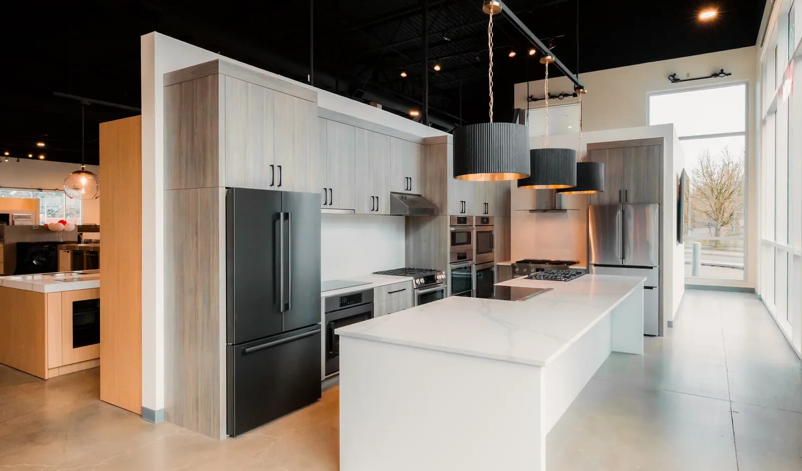 A kitchen appliance showroom under track lighting on an open black ceiling, fluted pendants over a white stone island