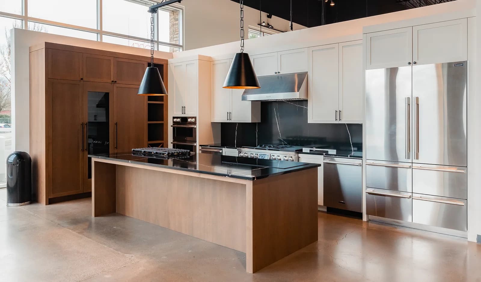 A showroom kitchen display with black cone pendants over a wood island, wall ovens and a range under a stainless hood