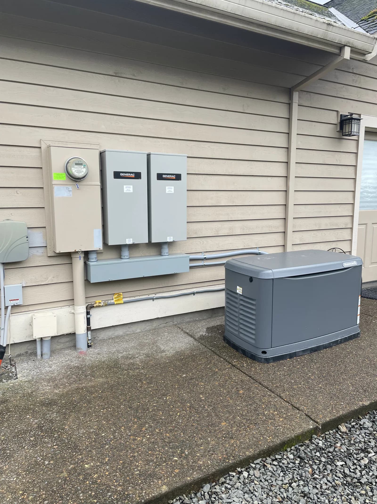 A meter and two transfer switches on a lap-sided wall, with a standby generator on the drive beside them