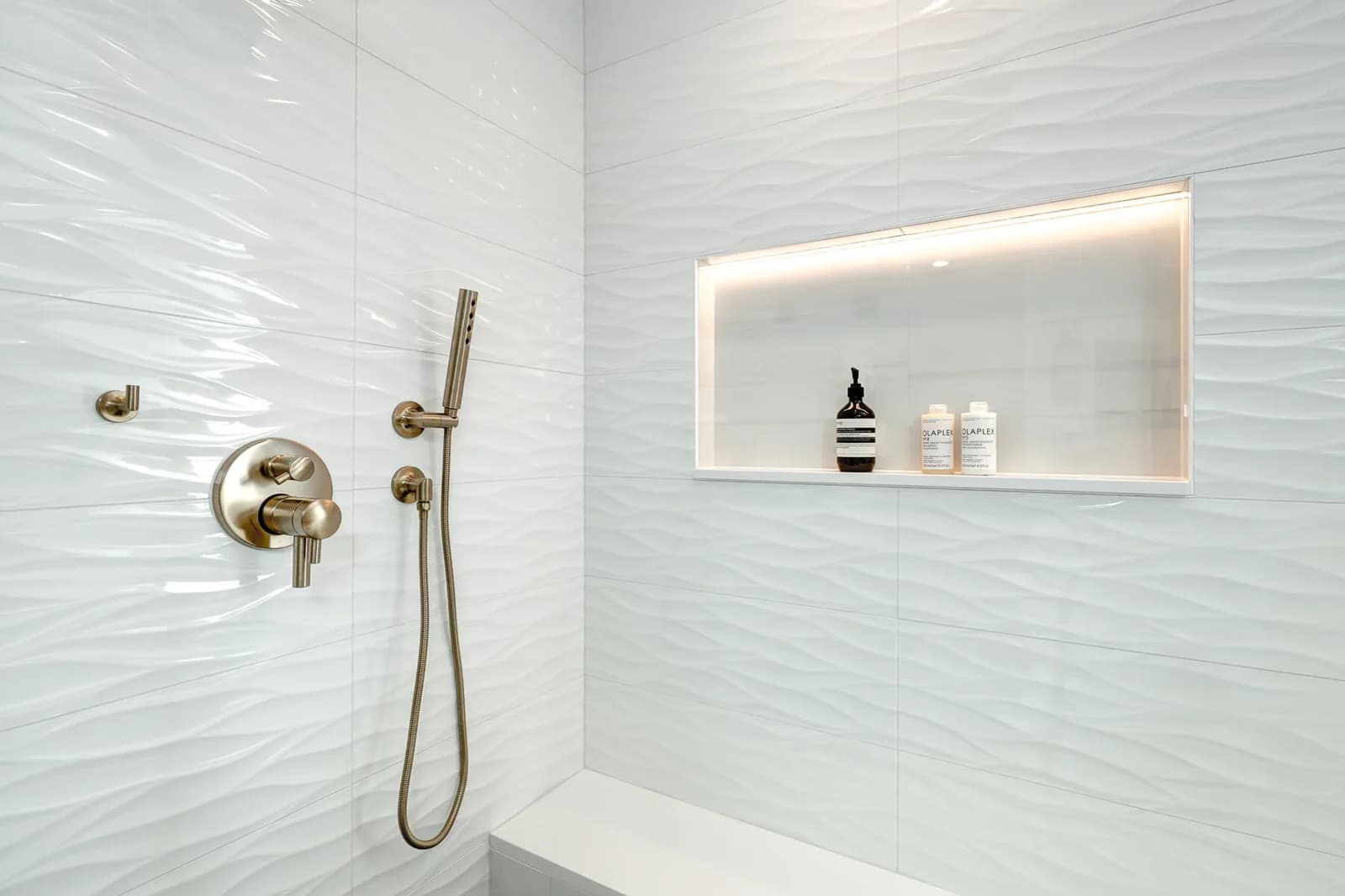 A lit recessed niche in a white tiled shower, beside a wall-mounted mixer and handset