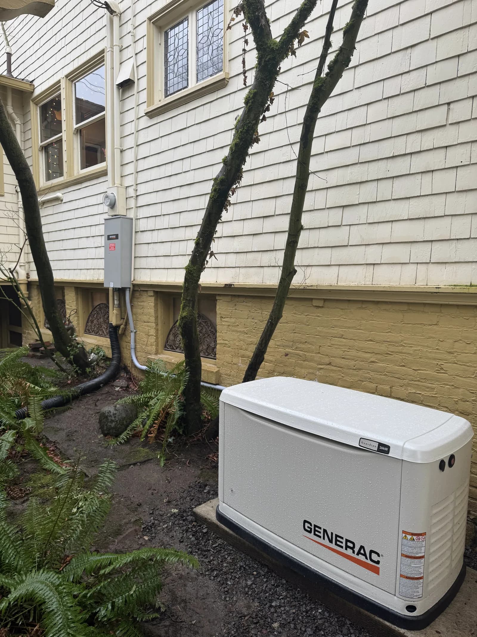 A white standby generator on a pad beside an older shingled house, transfer switch on the wall behind it