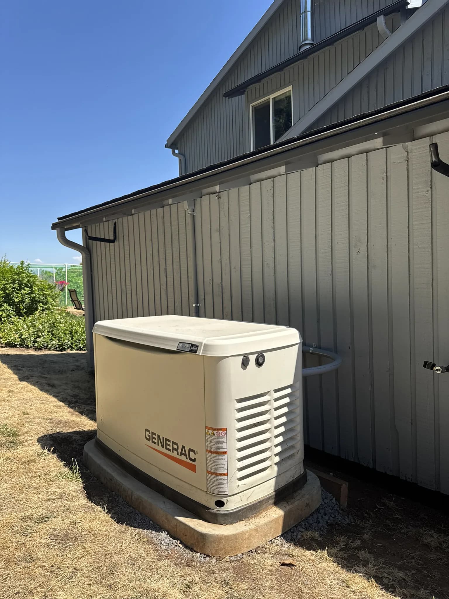 A Generac standby generator on a concrete pad beside a white board-and-batten house