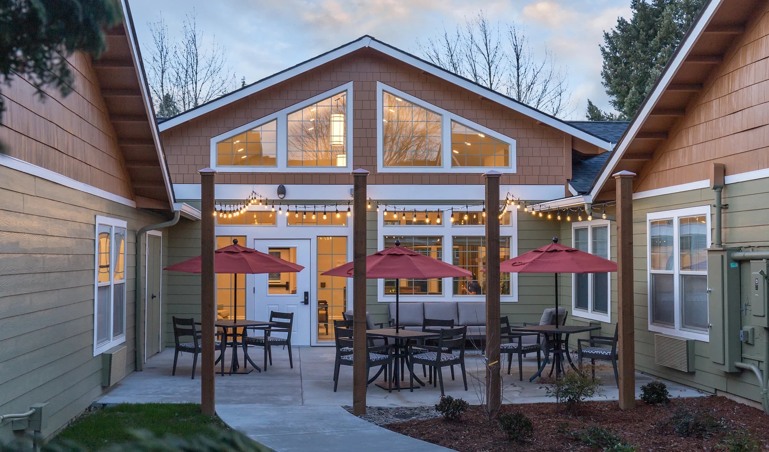 Lit courtyard at dusk, with string lights, umbrellas, and warm interior lighting through the windows