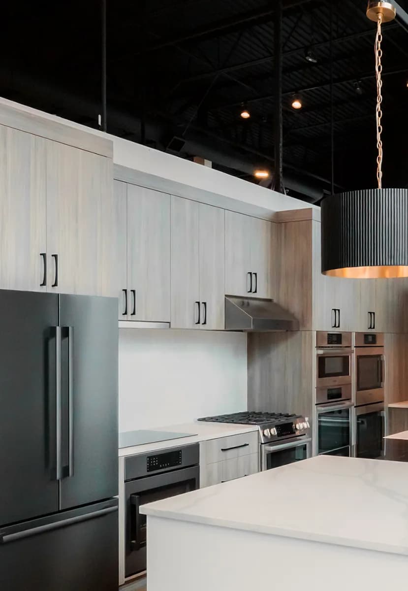 An appliance showroom lit by fluted pendants over a stone island, with track lighting and recessed downlights across a black open ceiling