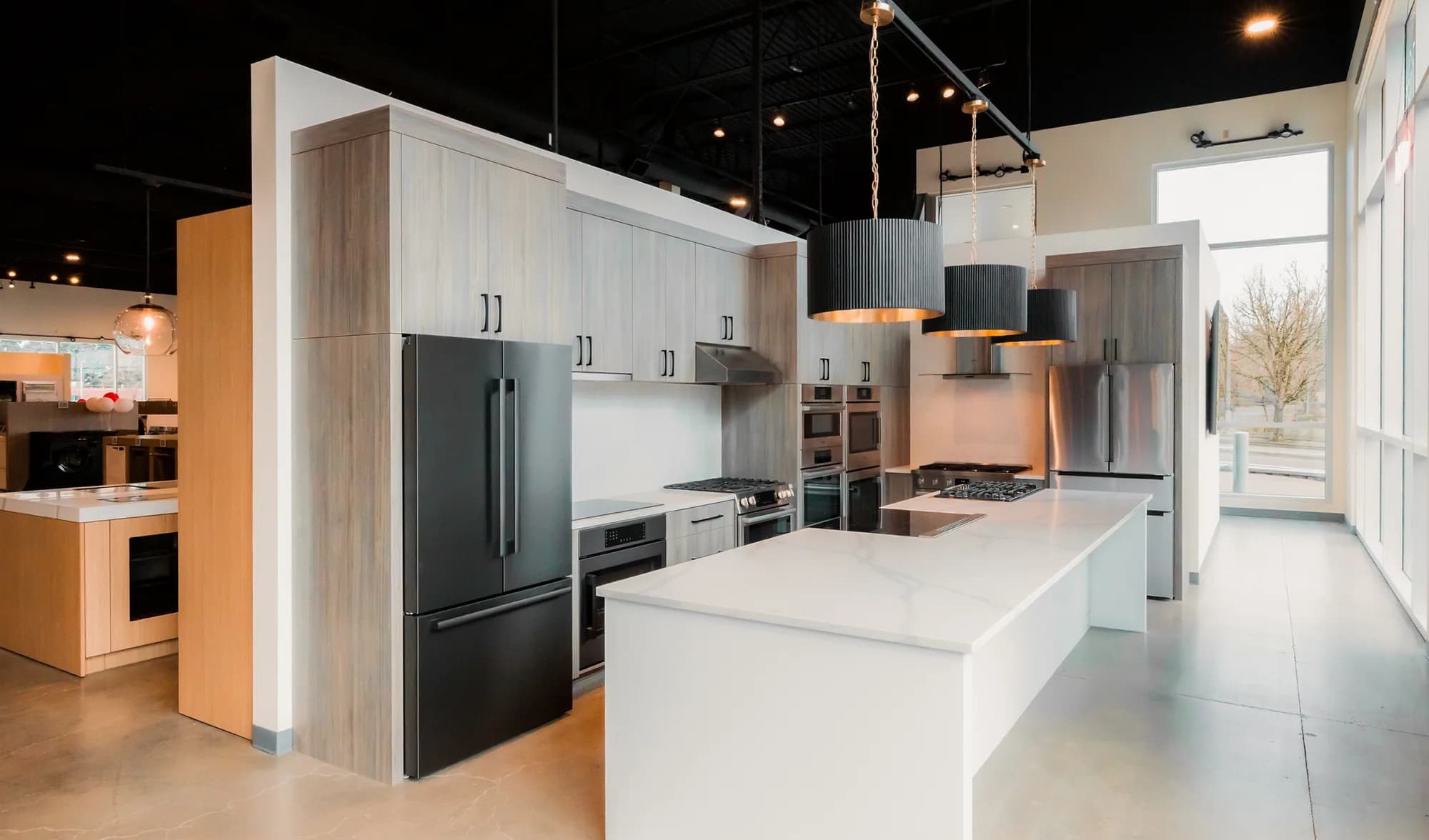 An appliance showroom lit by fluted pendants over a stone island, with track lighting and recessed downlights across a black open ceiling