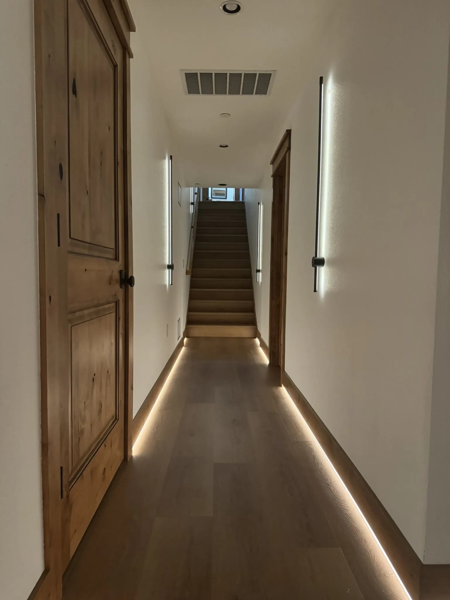 Rewired hallway lit by linear wall sconces, recessed cans, and a continuous LED strip along the baseboard, looking toward the stairs