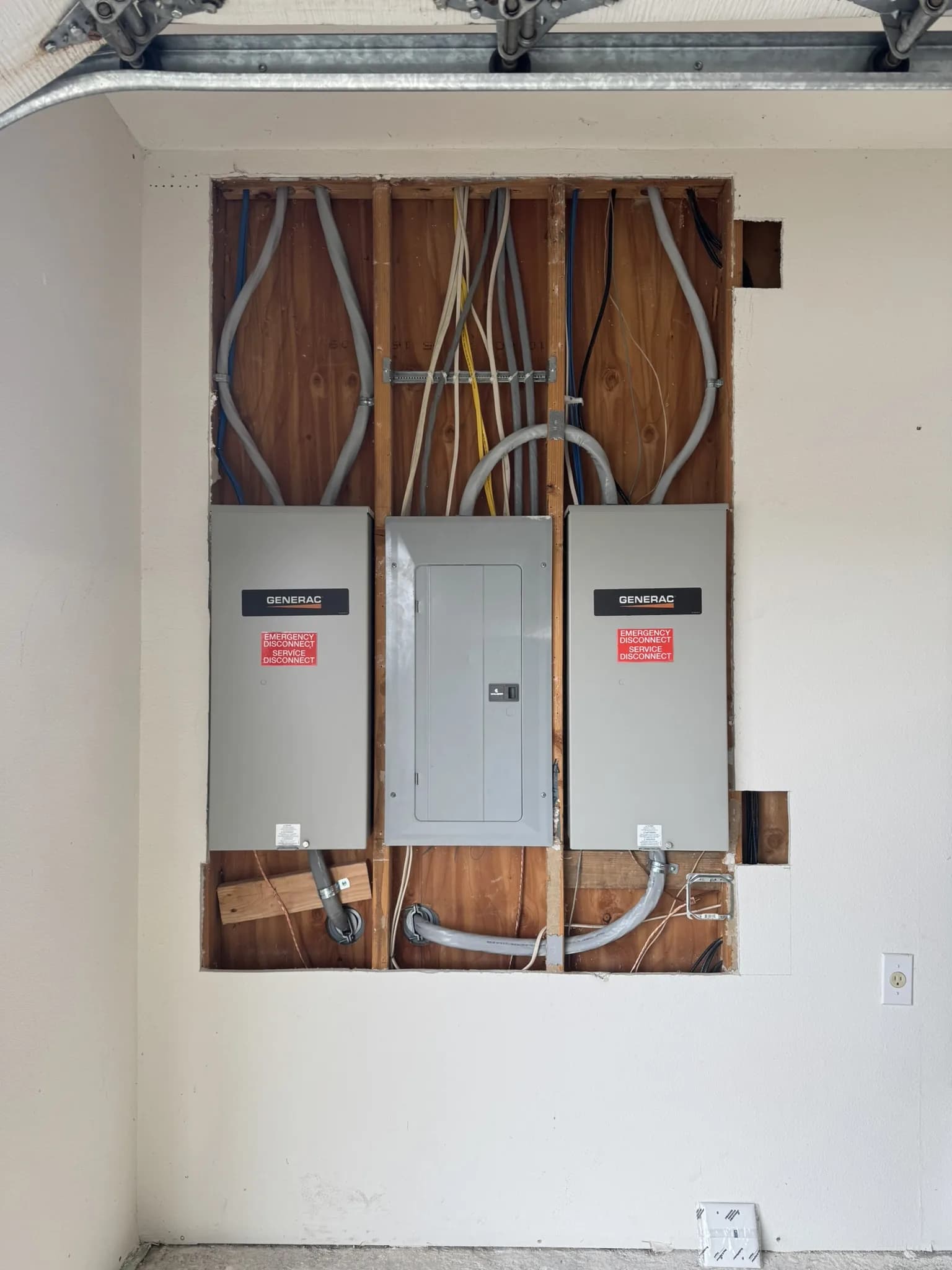 Garage wall opened to the studs around a new load center flanked by two Generac transfer switches, feeders and conduit run in to the panels